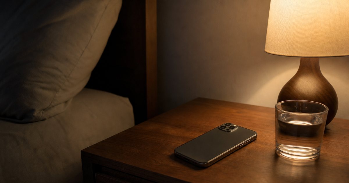 A smartphone lying face down on a wooden nightstand beside a switched-on bedside lamp and a glass of water, dark quiet bedroom at night, warm pool of light, soft shadows