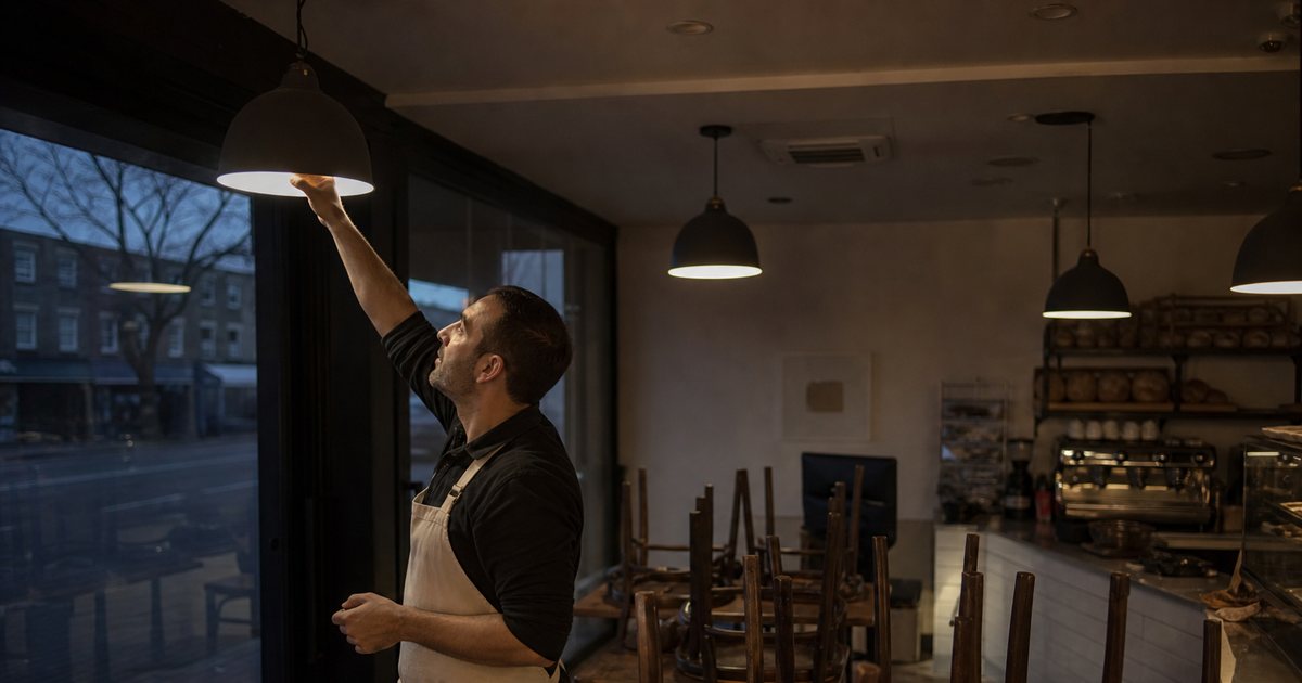 A bakery owner switching on the lights in a dim shop before dawn, chairs stacked upside down on tables, the street still dark through the front window